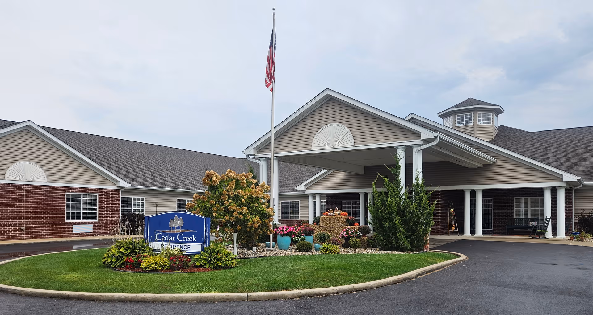 Exterior view of Cedar Creek Health Campus building with a covered entrance, landscaped garden with flowers and shrubs, an American flag on a flagpole, and a blue sign that reads 'Cedar Creek Residence'.