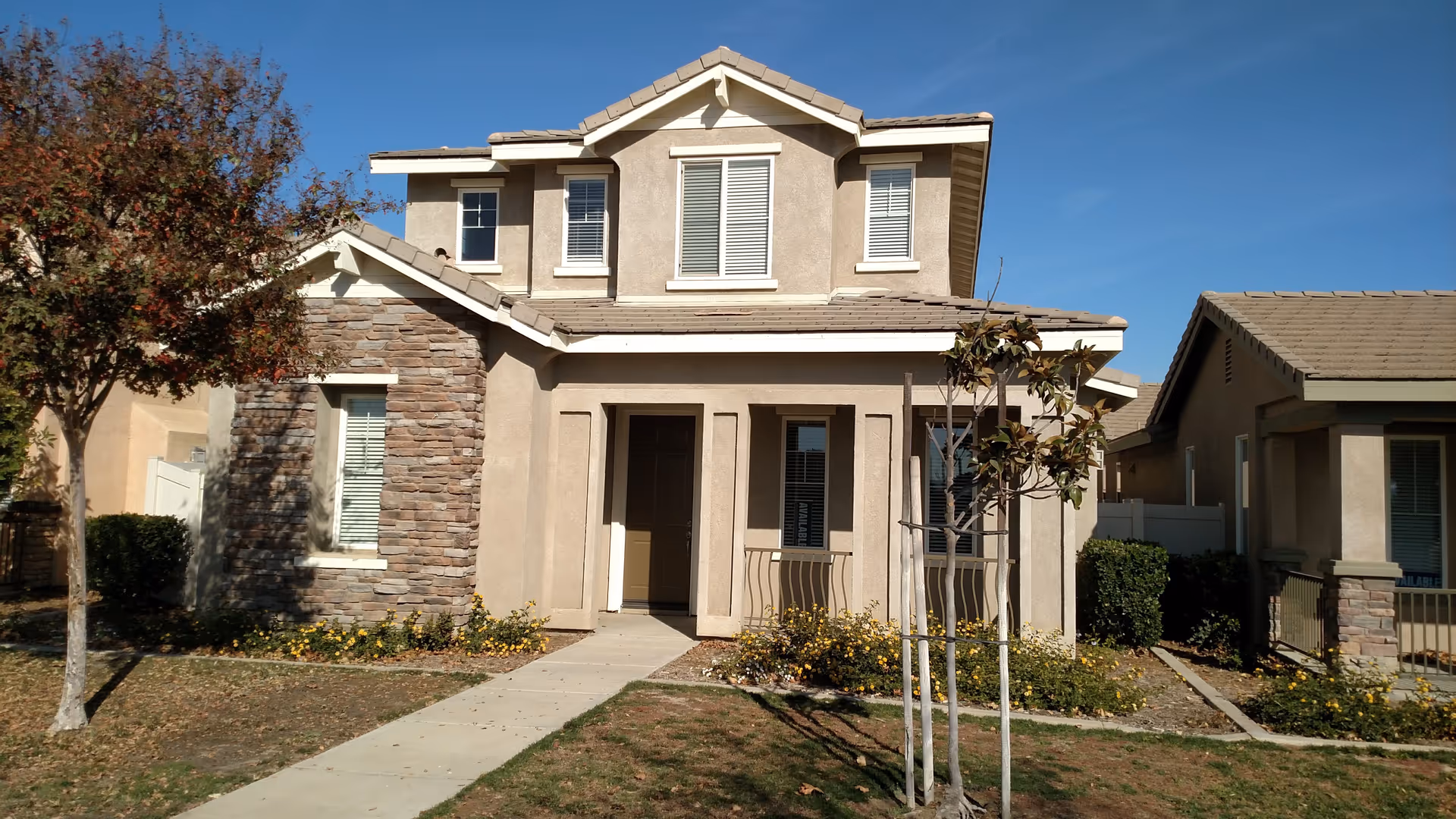 Front exterior view of a two-story residential building with beige stucco walls and stone accents. The house has a tiled roof, several windows with white shutters, a small front porch with a door, and a concrete walkway leading to the entrance. There are small trees and flower beds with yellow flowers in the front yard under a clear blue sky.