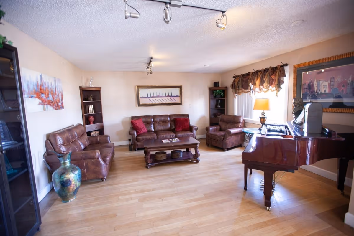 A cozy living room with wooden flooring featuring a brown leather sofa, two matching armchairs, a wooden coffee table with magazines, a grand piano, and decorative artwork on the walls. There are two tall bookshelves with decorative items and a large vase on the floor. A window with a curtain and a table lamp provide natural and artificial light.