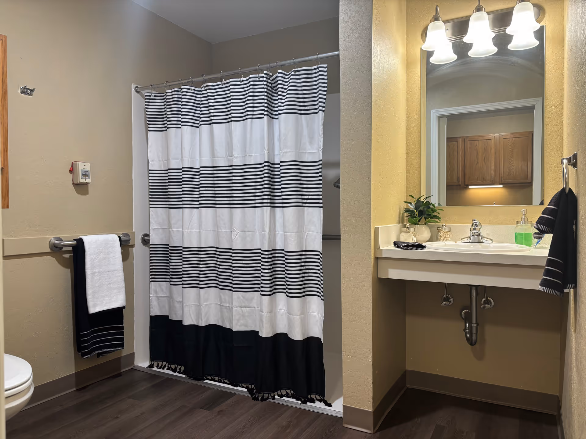 A bathroom with a shower area covered by a black and white striped shower curtain. There is a white toilet partially visible on the left, a towel rack with a white and black towel, a sink with a mirror above it, a small plant, soap dispenser, and a hand towel hanging on the right side. The floor is dark wood, and the walls are painted beige.