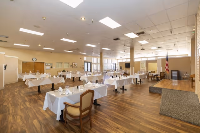 A spacious dining room with multiple tables covered in white tablecloths, each set with napkins and glassware. The room has wooden flooring, beige walls, and a small stage area with an American flag. Large windows allow natural light to fill the space.