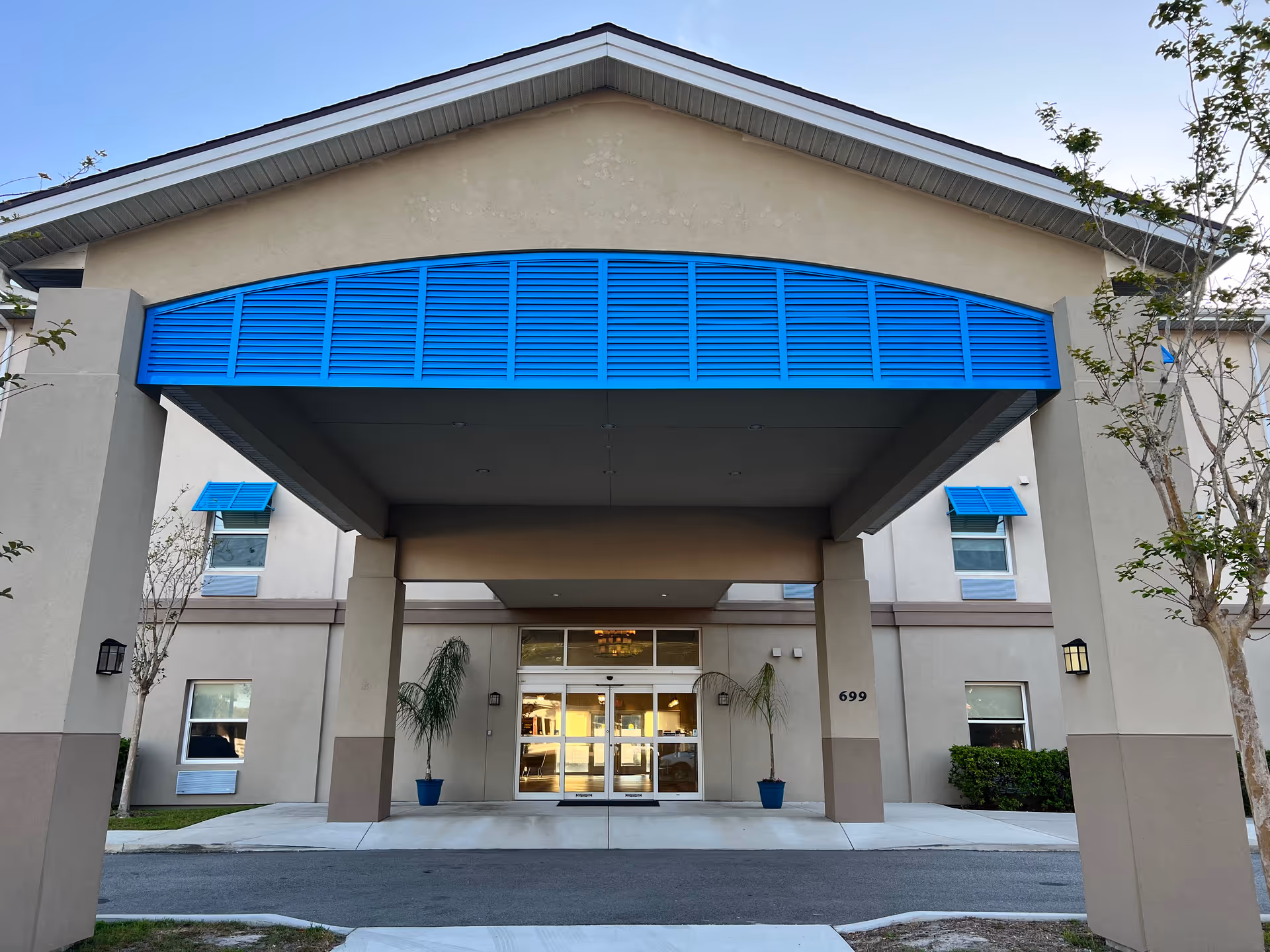 Front exterior view of a senior living facility entrance with a beige building and a large covered drop-off area featuring a blue accent above. Two potted plants flank the automatic glass doors, and the building number 699 is visible on one of the columns.