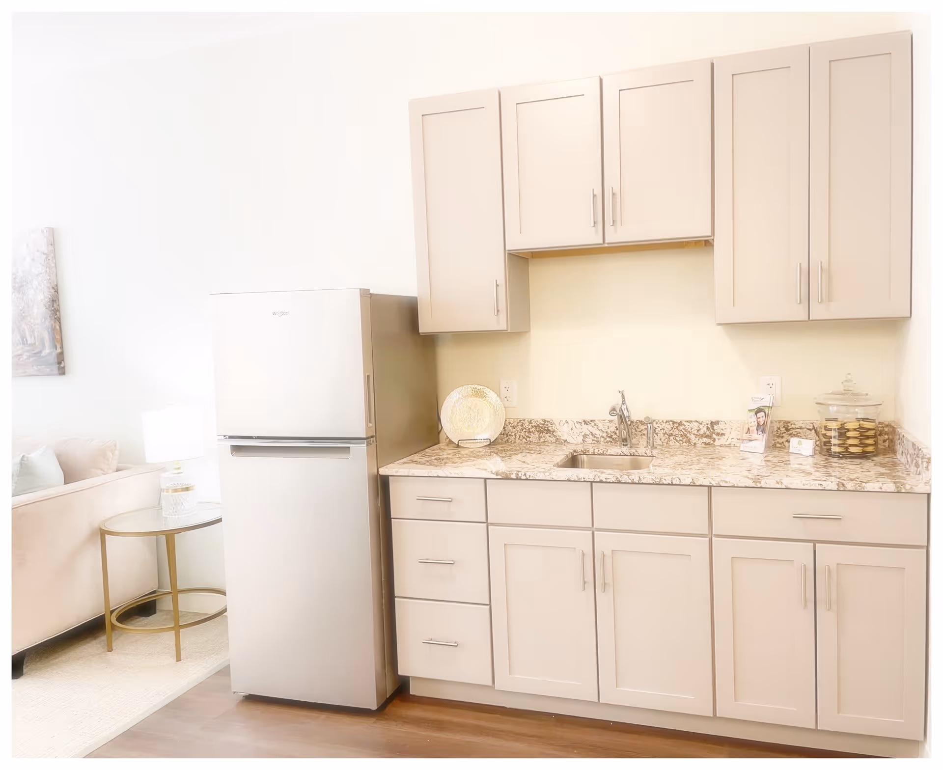 A bright kitchen area with beige cabinets, a marble countertop, a stainless steel refrigerator, and a small sink. To the left, part of a beige couch and a round glass side table with a lamp are visible, indicating an adjoining living space.