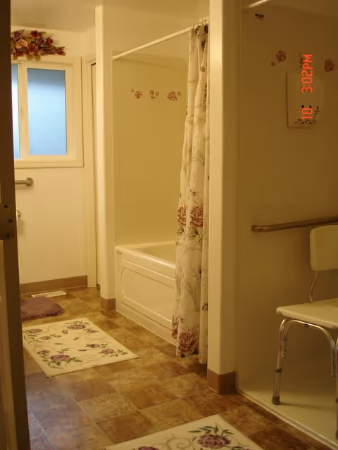 A bathroom with a bathtub partially covered by a floral shower curtain, floral rugs on the floor, a white chair with metal legs, a grab bar on the wall, a frosted window with floral decoration above it, and beige tiled flooring.