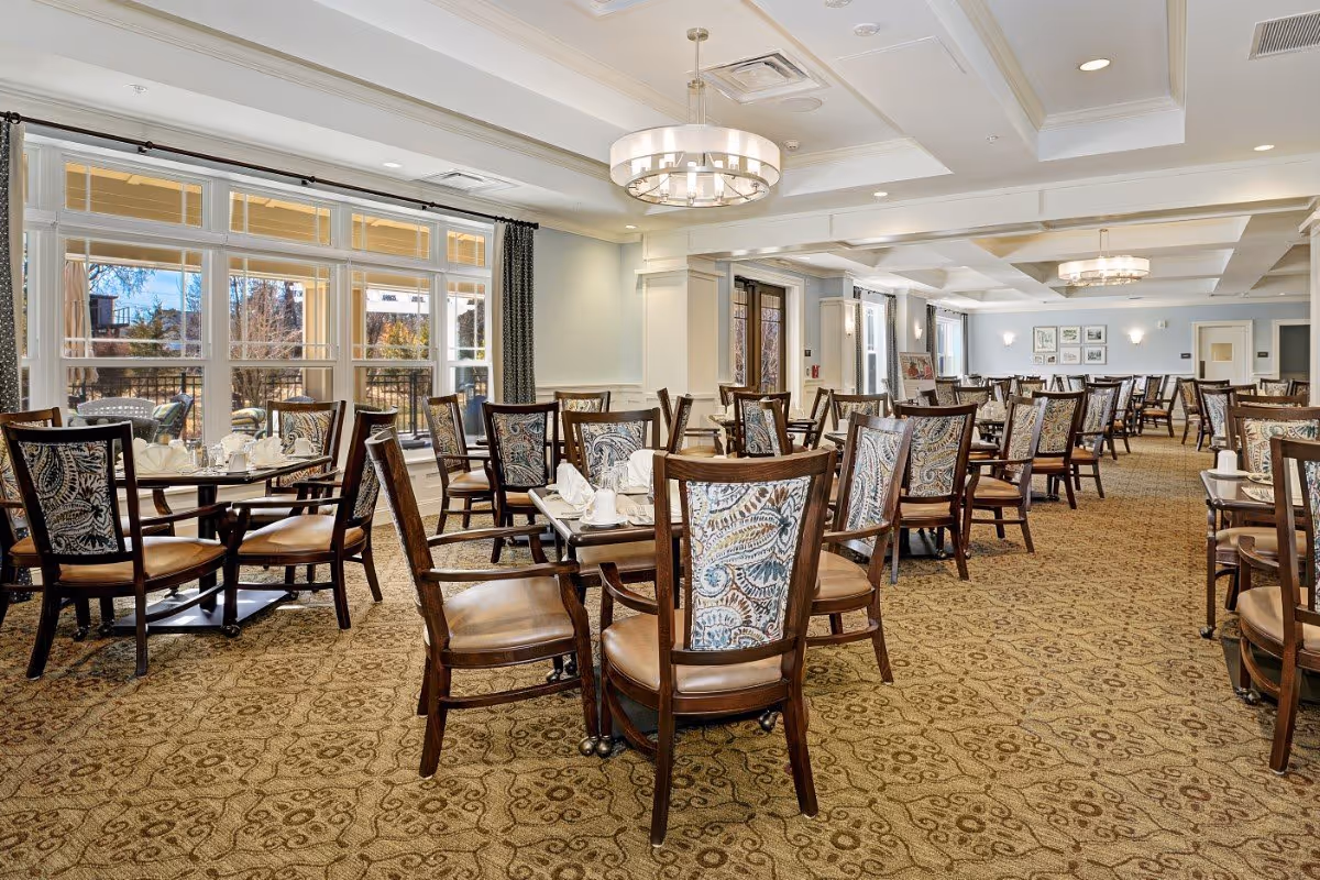 Large, well-lit dining room with multiple tables and patterned wooden chairs and place settings beside large windows.