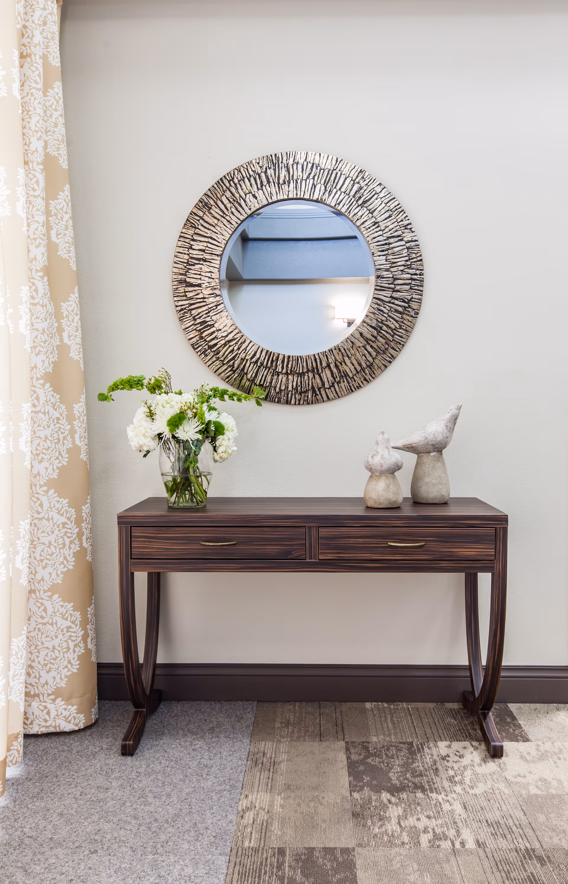 A wooden console table with two drawers against a light-colored wall. On the table, there is a glass vase with white and green flowers and two decorative bird sculptures. Above the table hangs a round mirror with a textured, patterned frame. To the left, beige curtains with white floral patterns are partially visible. The floor has a combination of carpet and patterned rug.