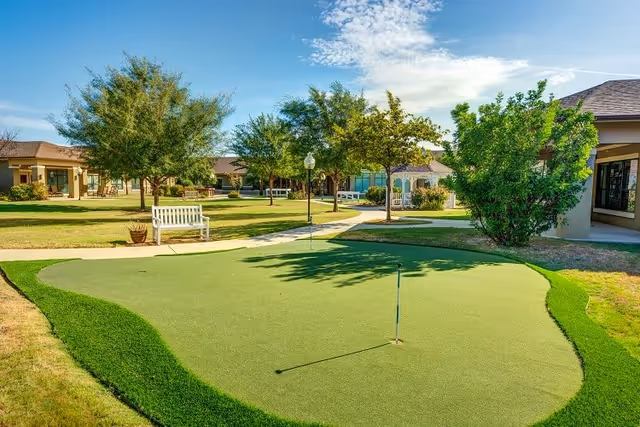 Outdoor putting green with two golf holes and flags, surrounded by a well-maintained lawn, trees, benches, and a gazebo in the background under a partly cloudy sky.