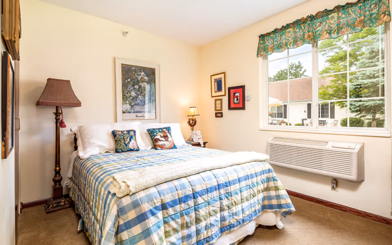 A cozy bedroom with a large bed covered in a blue and white checkered quilt and decorative pillows. There is a tall floor lamp on the left side and a small bedside table with a lamp and decorative items on the right. The room has a window with a floral valance, showing a view of trees and a building outside. The walls are decorated with framed artwork.