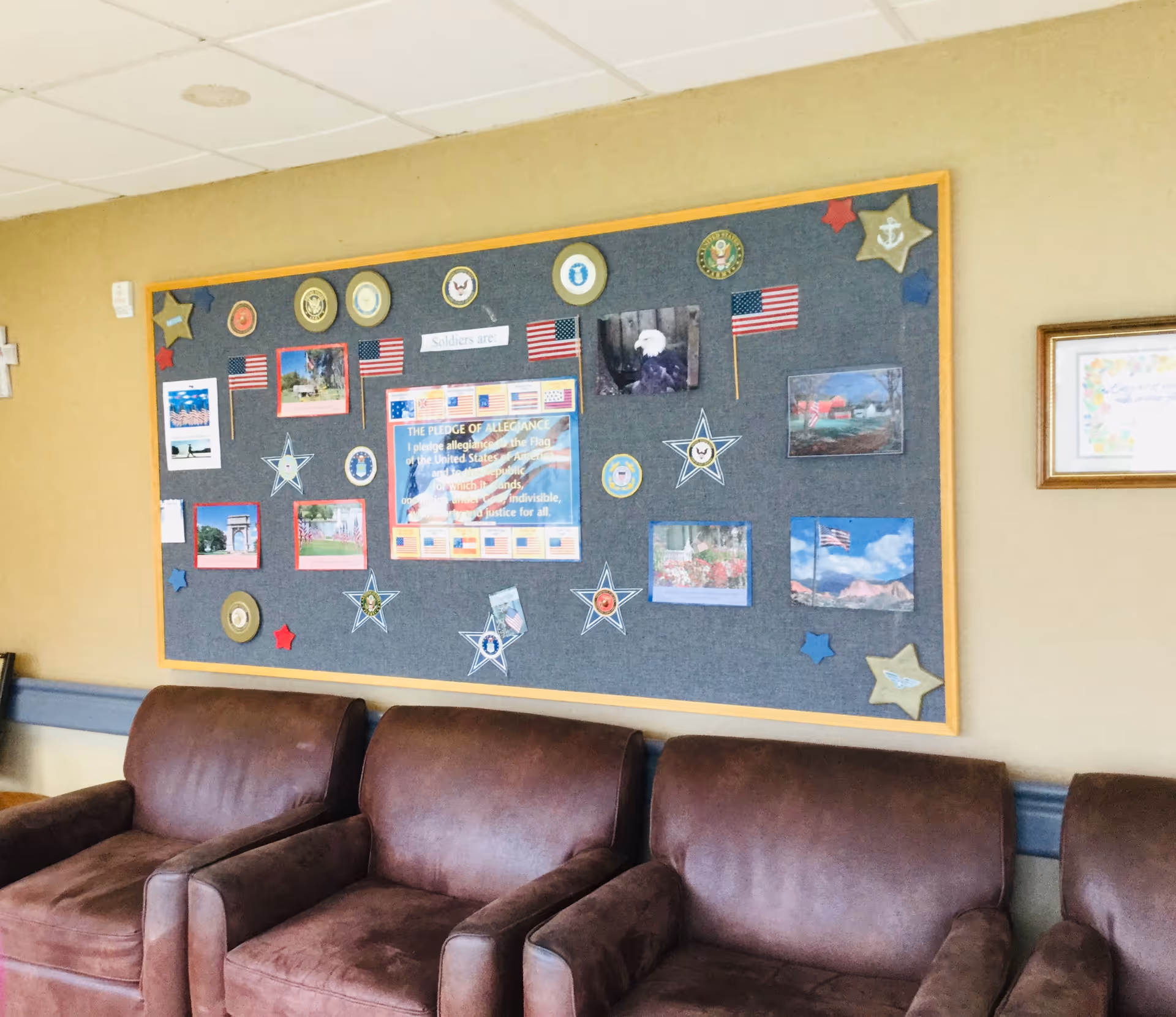 A seating area with three brown leather chairs in front of a wall decorated with a large bulletin board. The bulletin board features patriotic decorations including small American flags, stars, military insignias, and photos. The center of the board displays the Pledge of Allegiance.