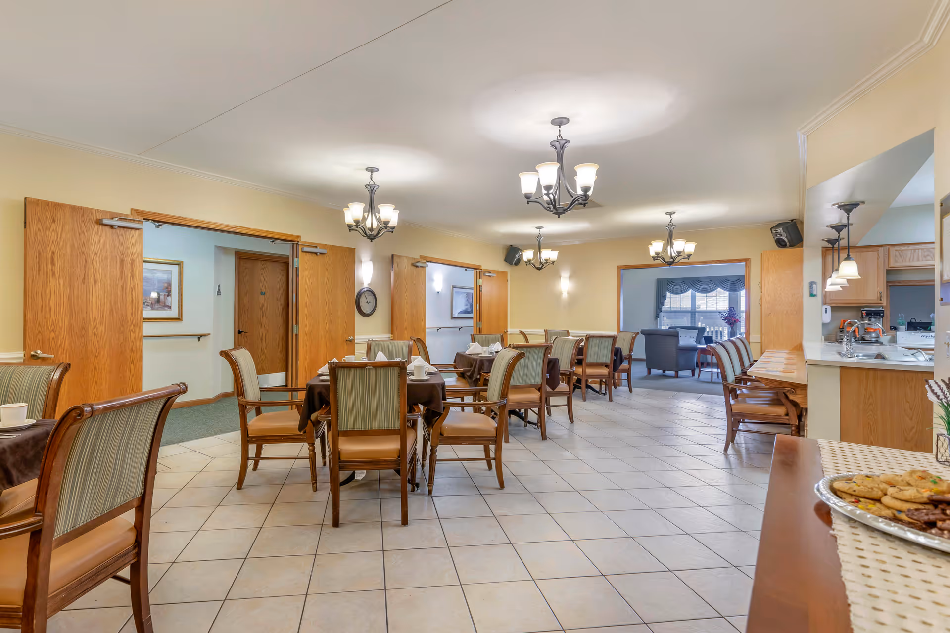 A dining room in Brookdale Midland with several tables covered in brown tablecloths and surrounded by wooden chairs with green striped upholstery. The room has beige walls, tiled floors, and multiple ceiling light fixtures. There are open wooden doors leading to other rooms, and a counter with a plate of cookies on the right side. In the background, a lounge area with armchairs and a window is visible.