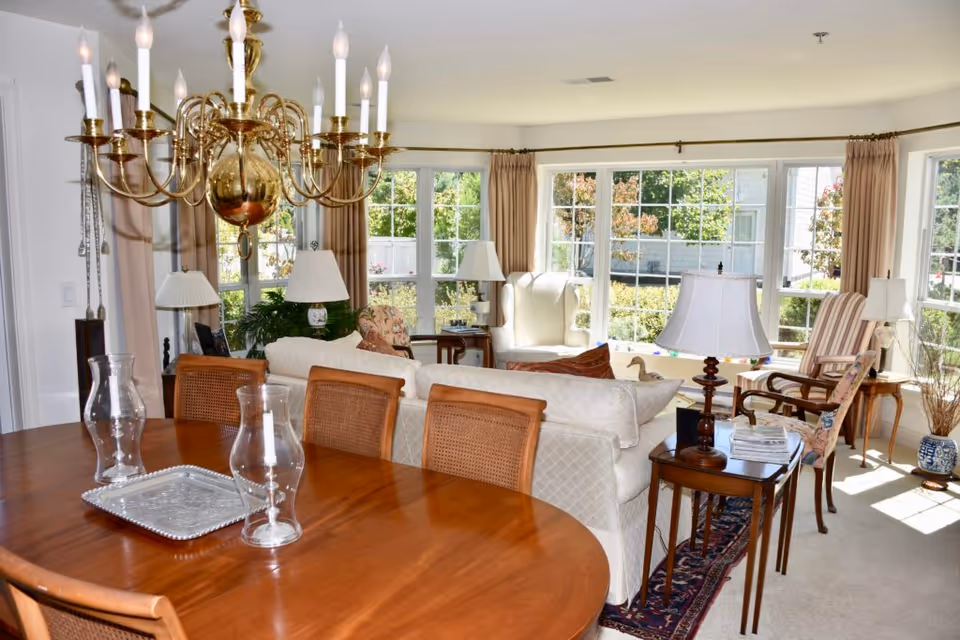 Bright sunlit living and dining area with a wooden dining table and chandelier in the foreground and upholstered sofas and chairs by large bay windows.