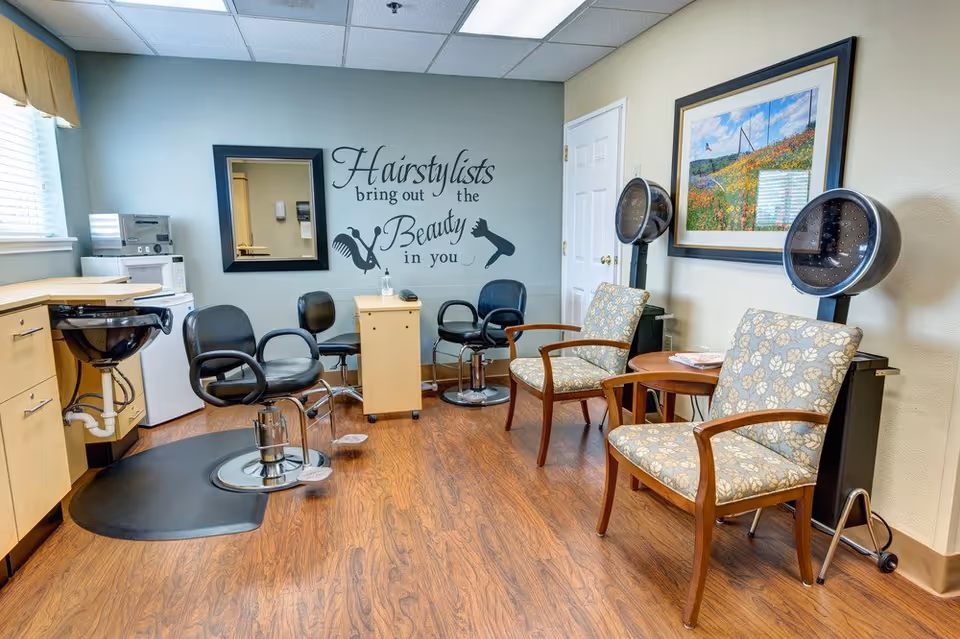 Interior of a hairstyling room with wooden flooring, two black salon chairs, two floral upholstered chairs, two hair drying stations, a black wash basin, a small wooden cabinet, a microwave, and a wall decal that reads 'Hairstylists bring out the Beauty in you'.
