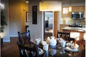 A dining area with a round glass table set for tea with white cups, saucers, and a teapot. Four dark wooden chairs surround the table. In the background, there is a kitchen with stainless steel appliances including a refrigerator, microwave, and stove. The room is warmly lit with pendant lights and has wooden flooring.
