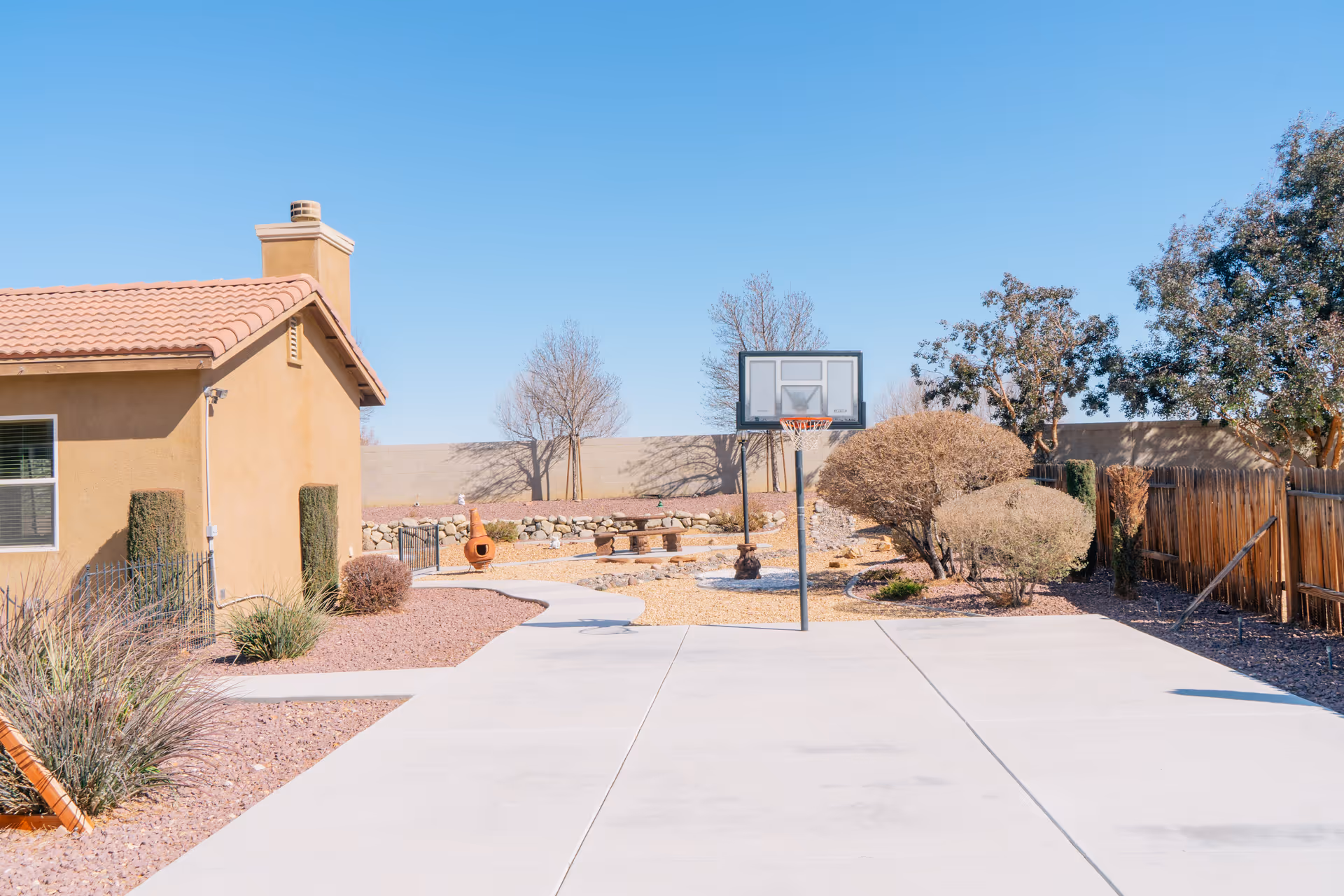 Outdoor area of a senior living facility with a basketball hoop on a concrete court, a tan building with a tiled roof on the left, landscaped bushes, trees, and a picnic table in the background under a clear blue sky.