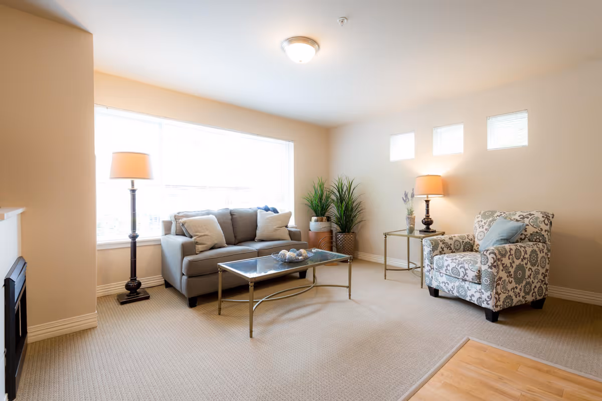 A bright living room with a large window letting in natural light. The room features a gray sofa with cushions, a patterned armchair with a blue pillow, a glass coffee table with decorative items, two floor lamps, and three small square windows on the wall. There are also two potted plants near the corner.