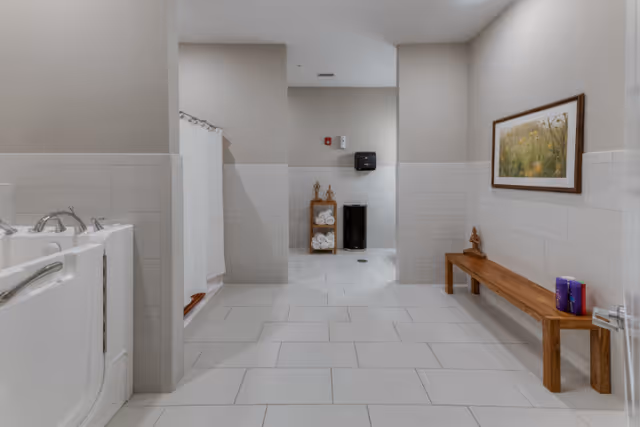 A clean and spacious bathroom area with a walk-in bathtub on the left, a shower with a white curtain in the center, and a wooden bench on the right. The walls are tiled halfway up with light-colored tiles, and there is a framed picture hanging above the bench. A small wooden shelf with rolled towels and decorative items is visible in the background, along with a black trash bin and a wall-mounted paper towel dispenser.