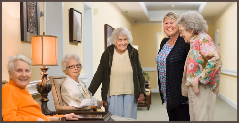 A group of four elderly women and one middle-aged woman standing and sitting in a well-lit hallway with light yellow walls and framed pictures. The elderly women are smiling and appear to be engaged in a friendly conversation.