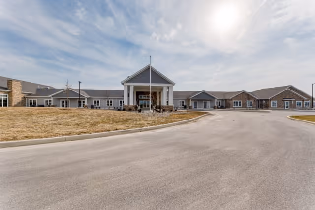 Front exterior of a single-story senior living facility with a covered entrance, circular driveway, and flagpole under a partly cloudy sky.