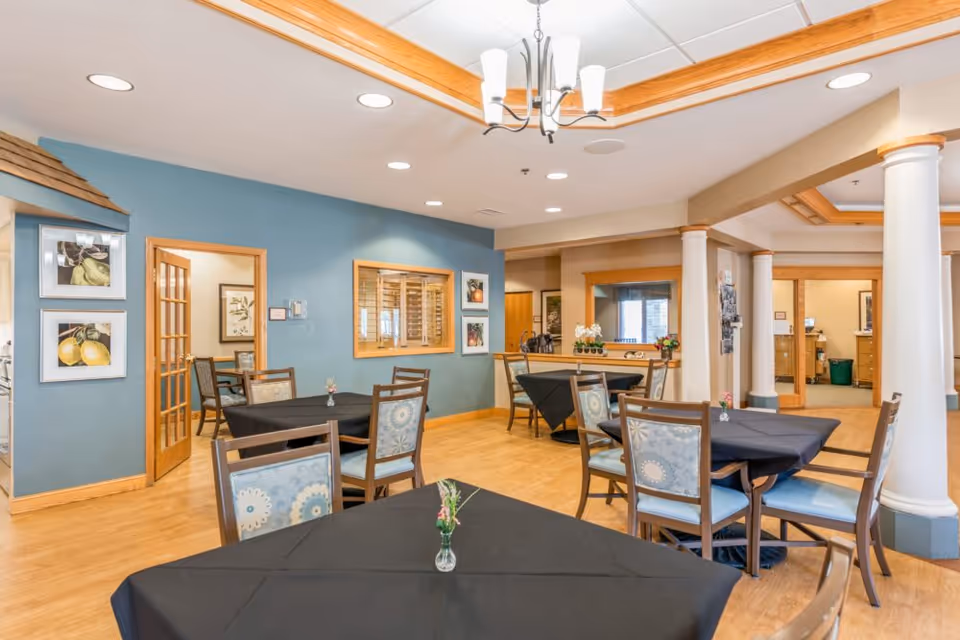 A bright dining area in a senior living facility with several tables covered in black tablecloths and chairs with patterned cushions. The room features wooden flooring, blue and beige walls, decorative columns, framed artwork, and a chandelier hanging from the ceiling.