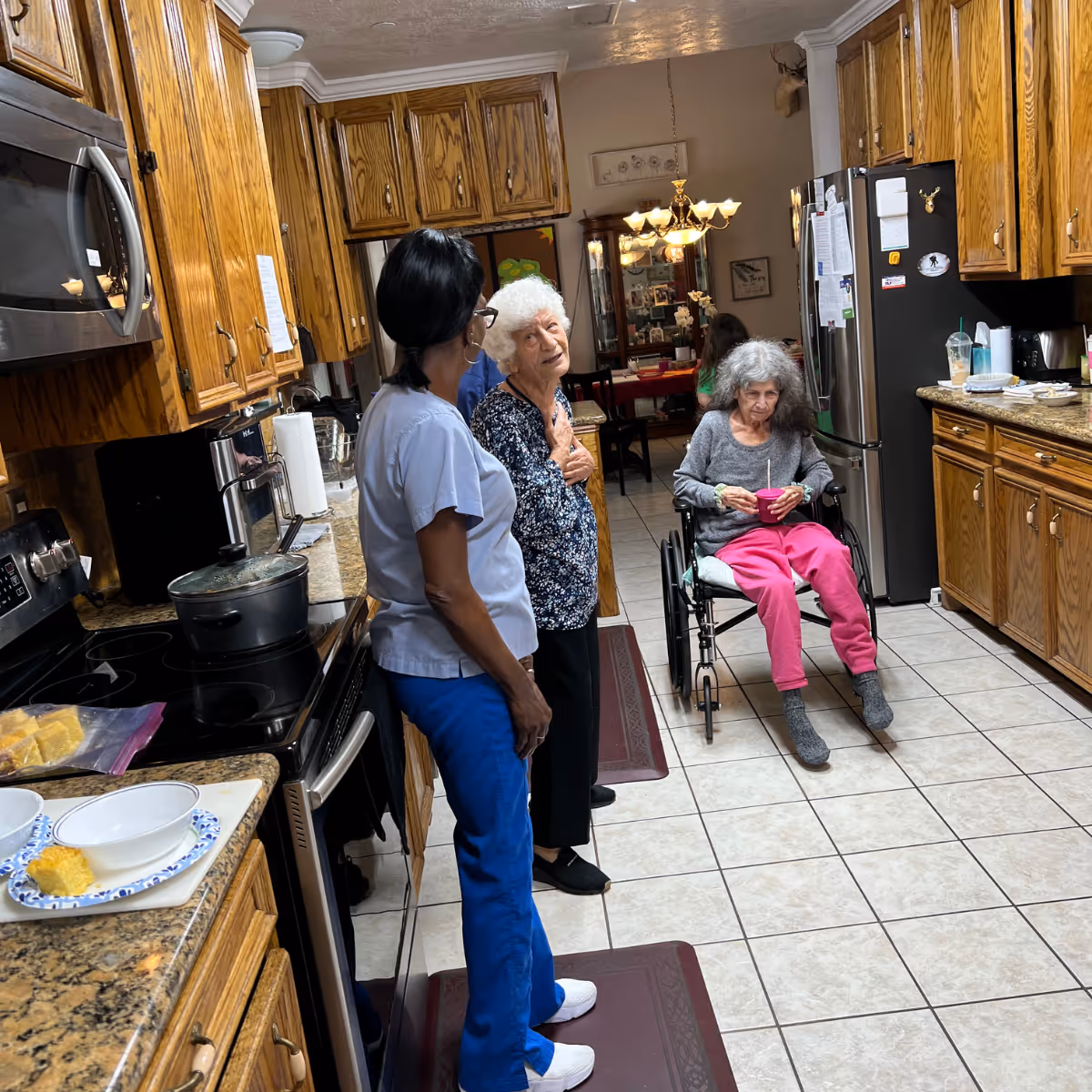 A kitchen scene in a senior care facility with wooden cabinets and granite countertops. Two elderly women are present, one standing and talking to a caregiver dressed in blue scrubs, and the other sitting in a wheelchair holding a pink bowl. The kitchen has tiled floors and a dining area visible in the background with a chandelier and a glass cabinet.