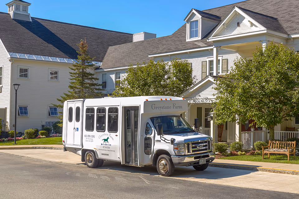 A white shuttle bus labeled Greystone Farm is parked in front of a large, white senior living facility building with a porch, trees, and a bench on a sunny day.