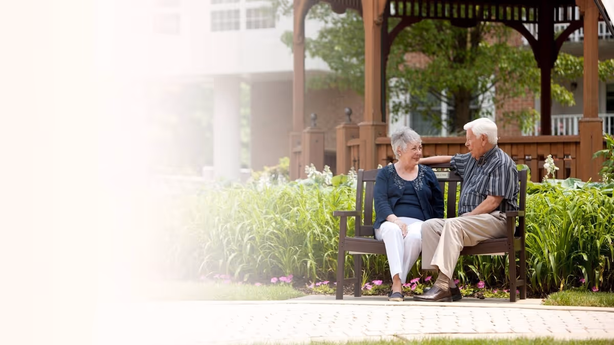 An elderly man and woman sitting on a bench in a garden area with green plants and flowers, engaged in conversation. Behind them is a wooden gazebo and part of a building is visible in the background.