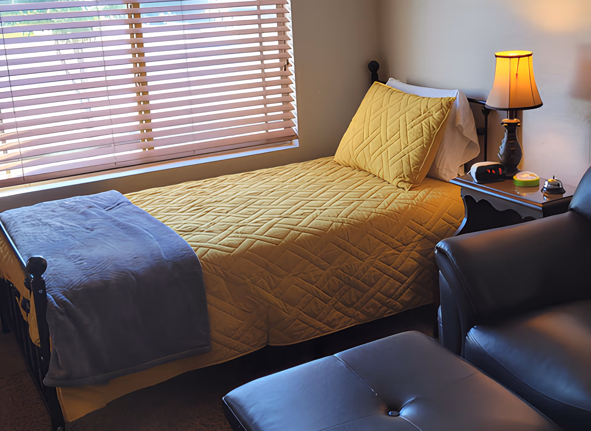 Sunlit bedroom with a single bed covered by a yellow quilt, a nightstand with a lamp and alarm clock, and a leather recliner.