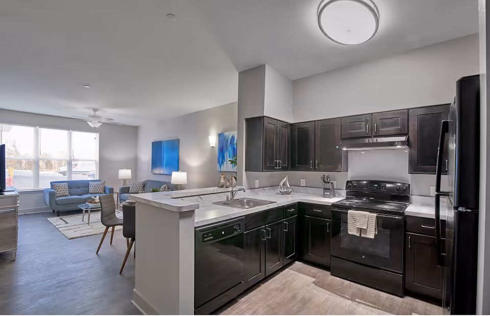 A modern open-concept kitchen and living room area in a senior living facility. The kitchen features dark wood cabinets, a black stove with a towel hanging on the handle, a black dishwasher, a black refrigerator, and a marble countertop with a sink. The living room has a blue sofa, a matching armchair, a coffee table, and two table lamps. Large windows allow natural light to fill the space, and blue artwork hangs on the walls.