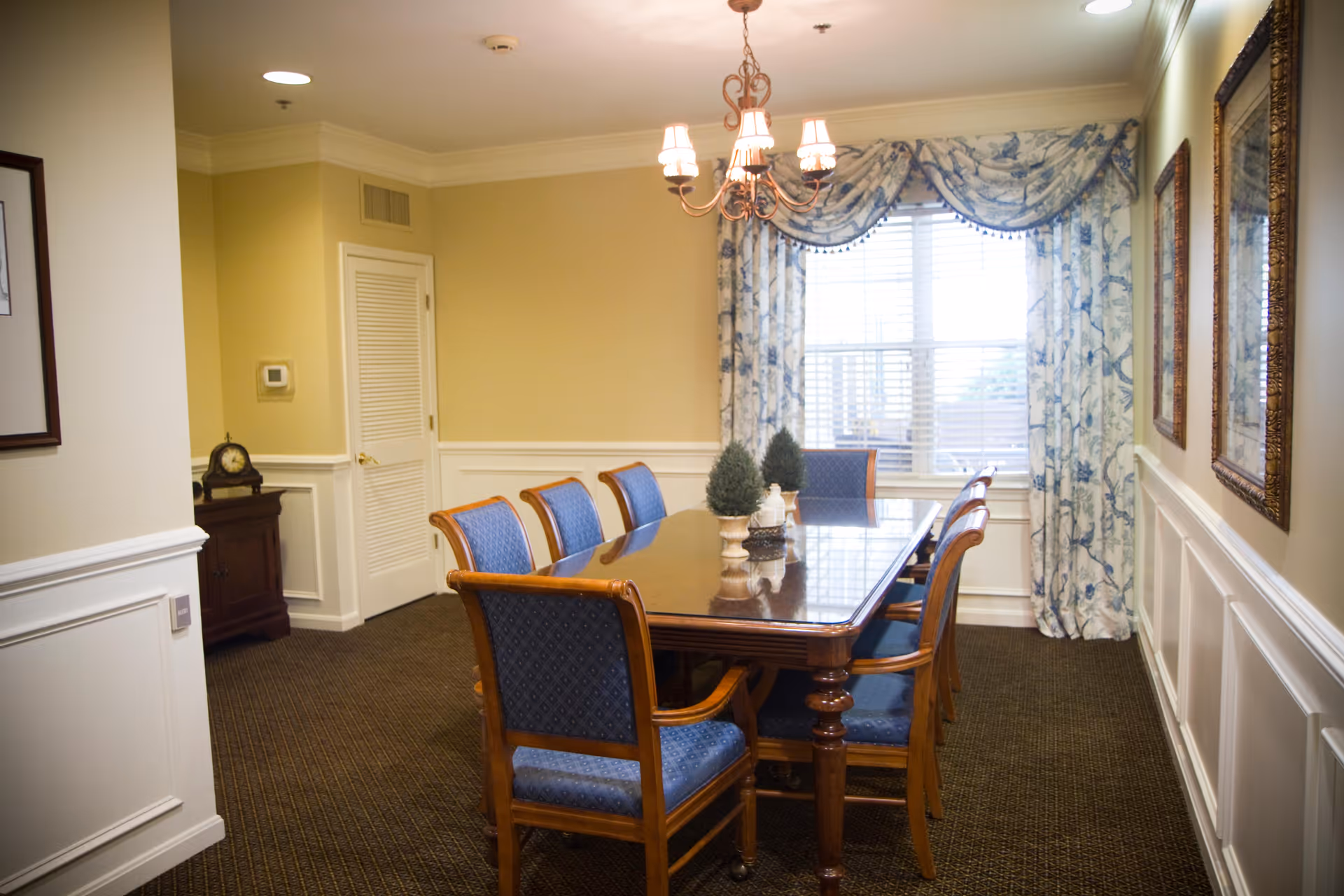 Formal dining room with a wooden table and blue upholstered chairs beneath a chandelier and a window with patterned curtains.