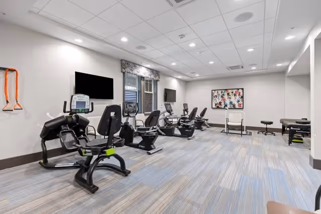 Well-lit indoor fitness room with several recumbent exercise bikes, wall-mounted TVs, and a treatment table.