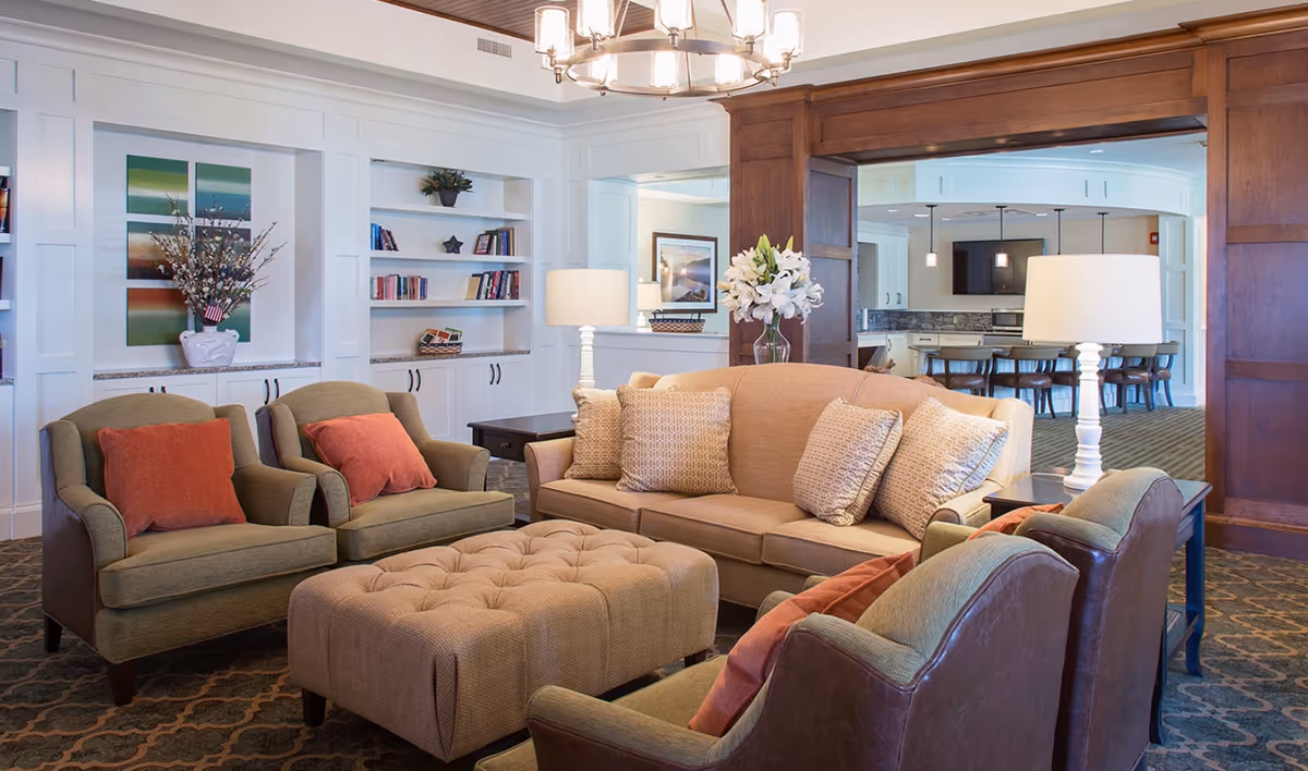 A cozy living room area with a beige sofa, four armchairs with orange cushions, and a large tufted ottoman in the center. The room features built-in white shelves with books and decorative items, two table lamps, a vase with white flowers, and a wooden archway leading to a kitchen or dining area with bar stools and pendant lights.