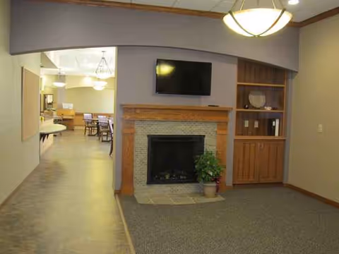 Common lounge area with a fireplace, mounted TV, wooden shelves, and seating in a senior facility.