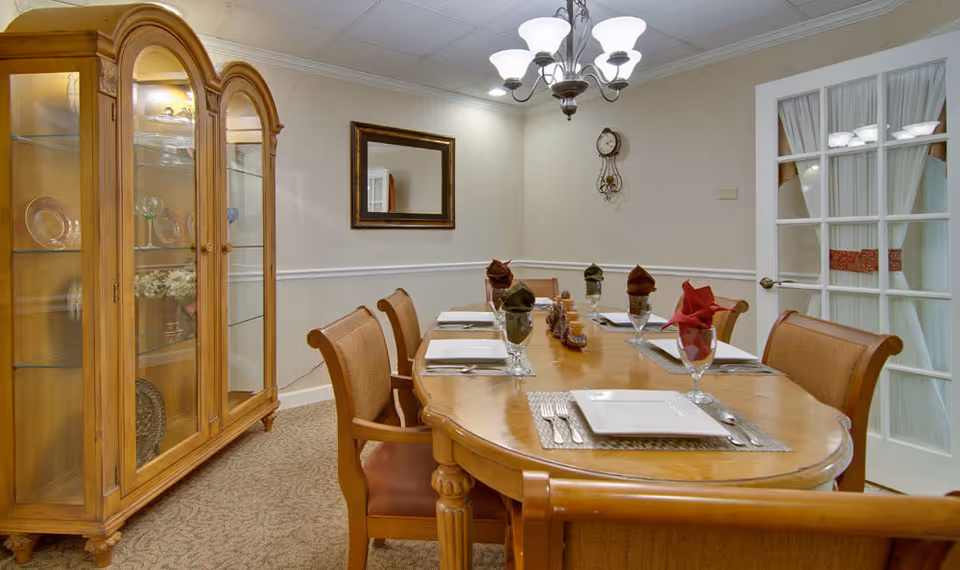 A dining room with a wooden dining table set for six people. The table has placemats, square white plates, silverware, glasses with folded napkins in red and green, and a centerpiece. There are six wooden chairs with cushioned seats around the table. On the left side, there is a wooden china cabinet with glass doors displaying plates and decorative items. The room has beige walls with a white chair rail, a wall clock, a rectangular mirror, and a chandelier hanging from the ceiling. A door with glass panes and white curtains is visible on the right side.