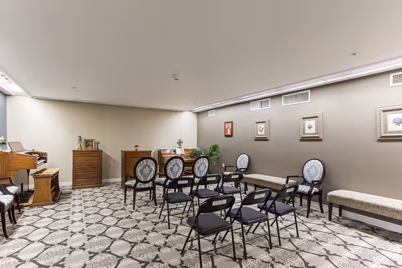 A small room with patterned tile flooring, several folding chairs arranged in rows facing two wooden organs and a piano. The walls are decorated with framed pictures and there are upholstered chairs and benches along the right wall.