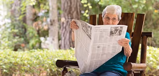 An elderly woman with white hair and glasses sitting outdoors on a wooden rocking chair, reading a newspaper. The background shows greenery and trees, suggesting a peaceful garden or patio setting.