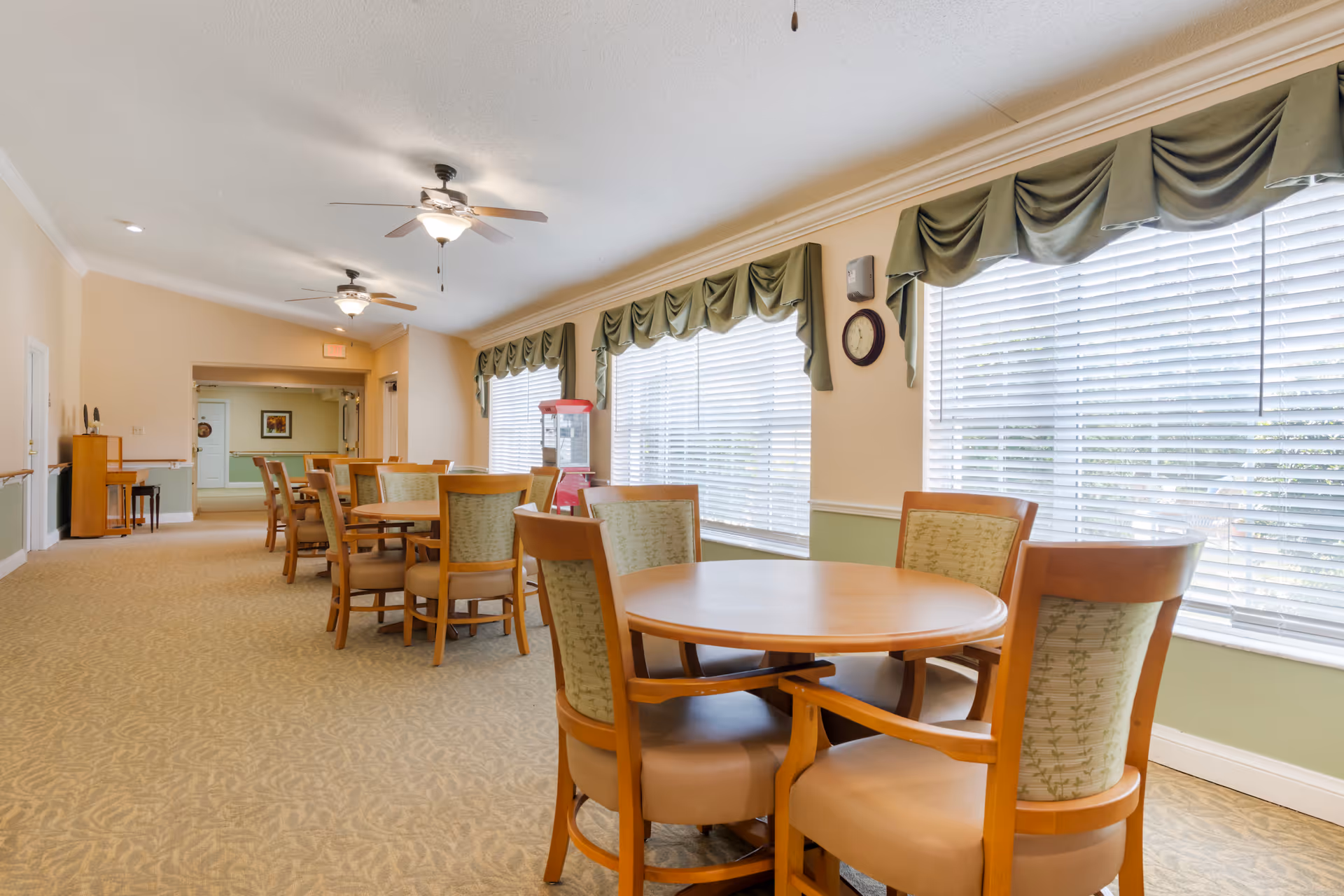 A bright dining area with round wooden tables and cushioned chairs arranged in rows along large windows with green valances. Ceiling fans with lights hang from the white ceiling, and a popcorn machine is visible near the windows. The room has beige walls with green wainscoting and carpeted floors.