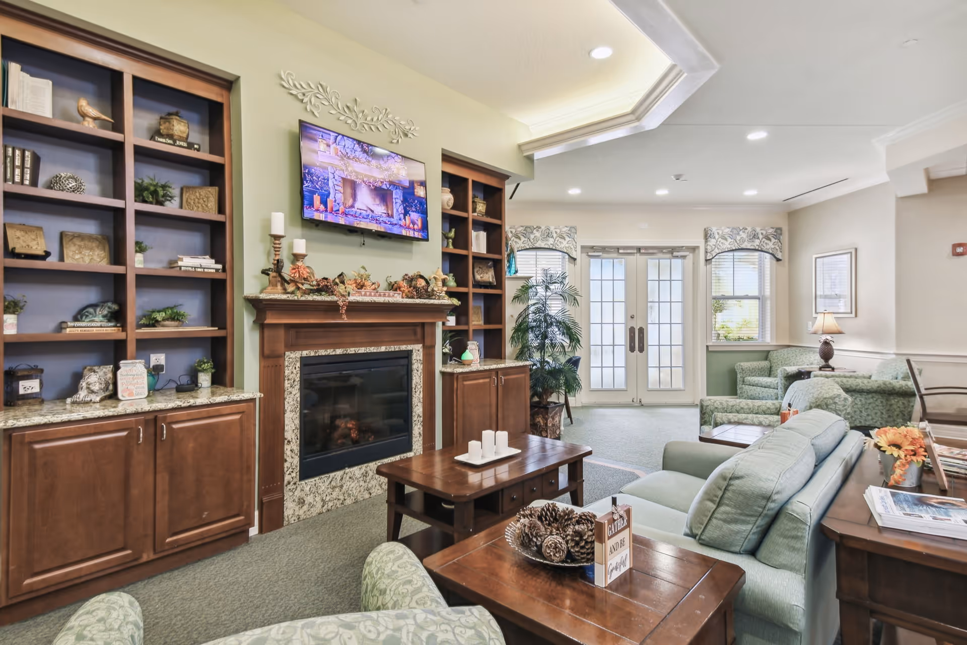 A cozy living room area in a senior living facility featuring a fireplace with a mounted TV above it, built-in wooden shelves on either side, comfortable seating including armchairs and sofas, a coffee table with decorative pinecones, and large windows with floral valances letting in natural light.