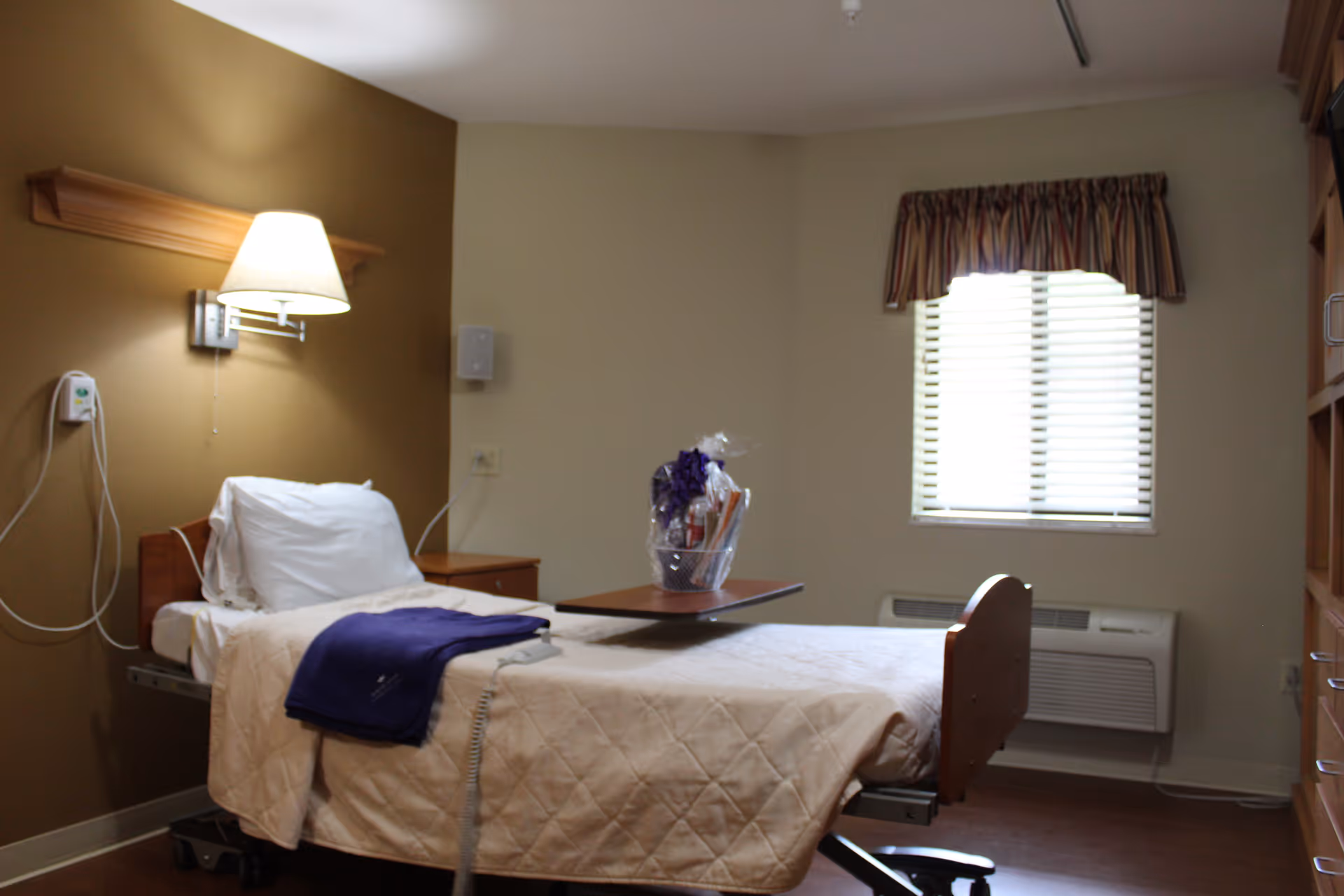 A single bed in a senior living facility room with beige bedding and a purple blanket folded on top. There is a wall-mounted lamp above the bed, a small bedside table, and a window with blinds and a valance. A gift basket is placed on an overbed table. The room has neutral-colored walls and a wooden cabinet on the right side.