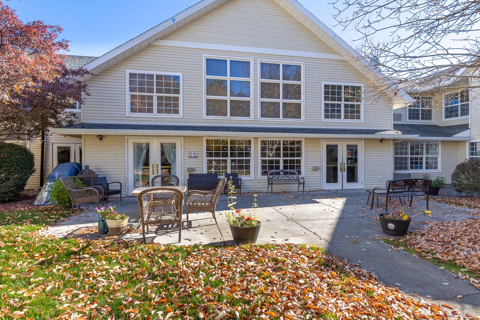 Outdoor patio area of McKay Creek Assisted Living with several wicker chairs and benches arranged on a concrete surface. There are potted plants with flowers placed around the patio. The building has beige siding with large windows and two sets of double glass doors. Autumn leaves cover the grass and parts of the patio, and trees with fall foliage are visible.