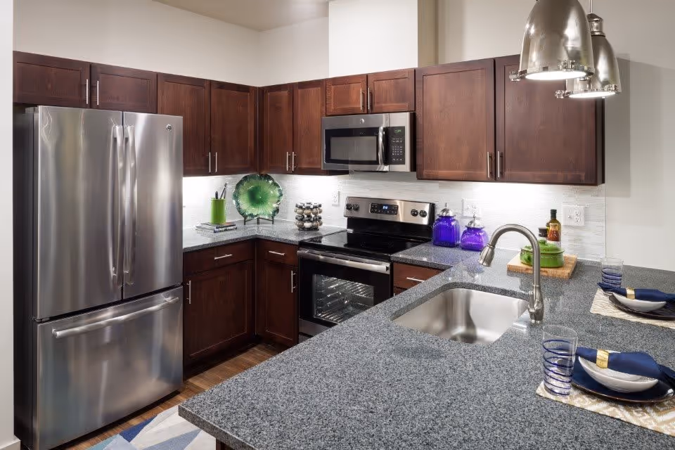 Modern kitchen with dark wooden cabinets, stainless steel refrigerator, oven, microwave, and a granite countertop with a built-in sink. The countertop has place settings with blue glasses, white plates, and navy napkins. Decorative items include a green glass plate, purple jars, and a small cutting board with bottles.