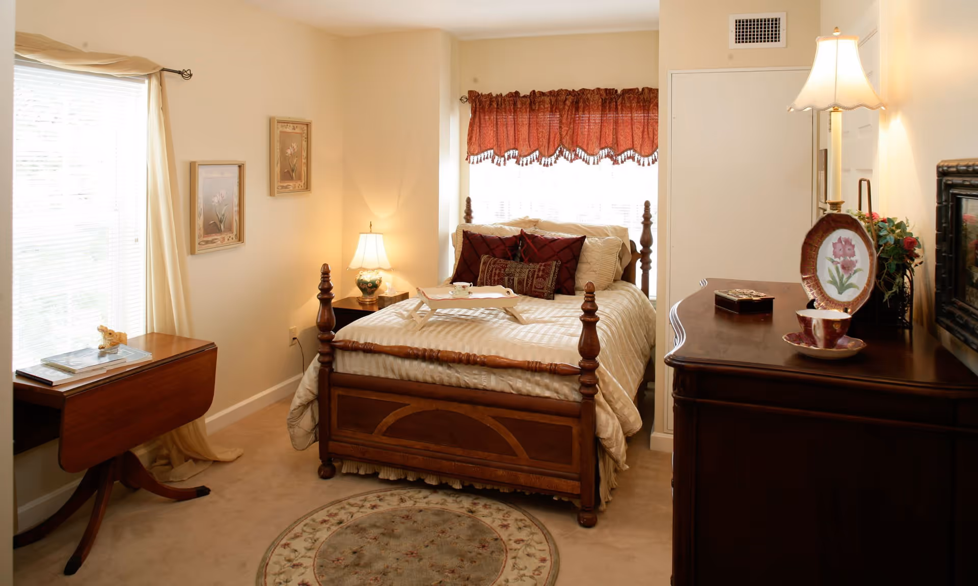 A cozy bedroom with a wooden bed featuring beige and burgundy bedding, a small wooden side table with a lamp, a wooden dresser with a decorative plate and teacup, a window with red valance curtains, and a small round rug on the carpeted floor.