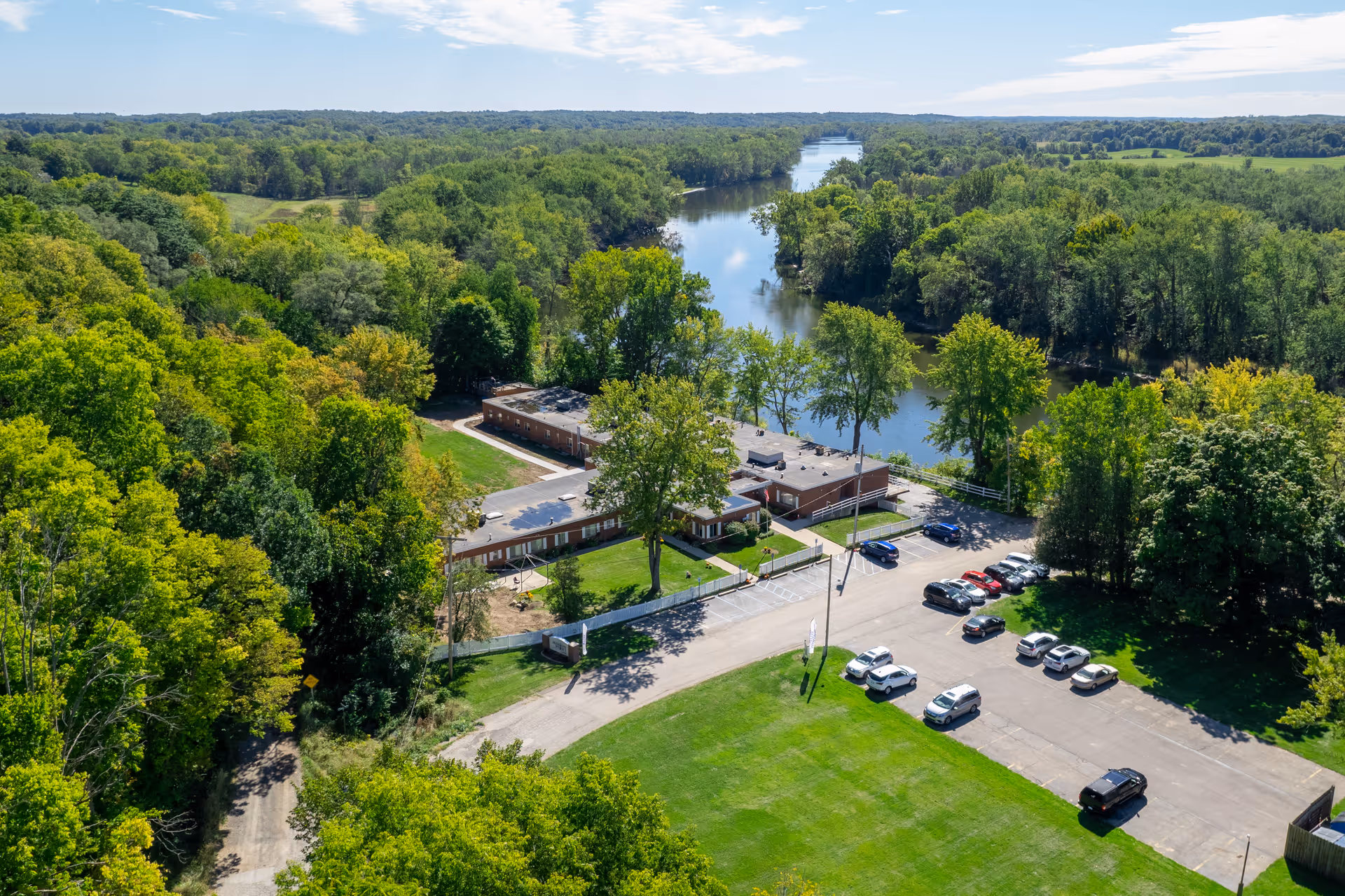 Aerial view of Intersect Healthcare of Lamont facility surrounded by dense green trees and situated next to a river. The building is a single-story structure with a parking lot containing several cars. The landscape includes a large grassy area and a winding road leading to the facility.