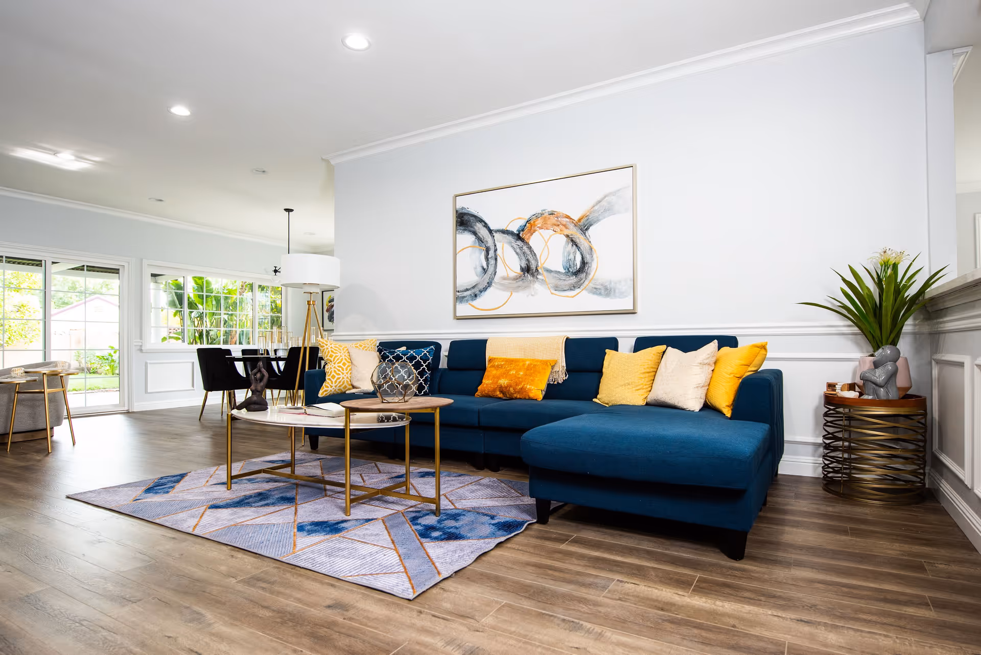 A modern living room with a blue sectional sofa adorned with yellow and beige pillows. A geometric patterned rug lies underneath two round coffee tables with gold legs. A large abstract painting with circular designs in black, gray, and orange hangs on the white wall above the sofa. To the right, there is a side table with a plant and decorative items. In the background, a dining area with a black dining table and chairs is visible near large windows and a sliding glass door that leads outside.