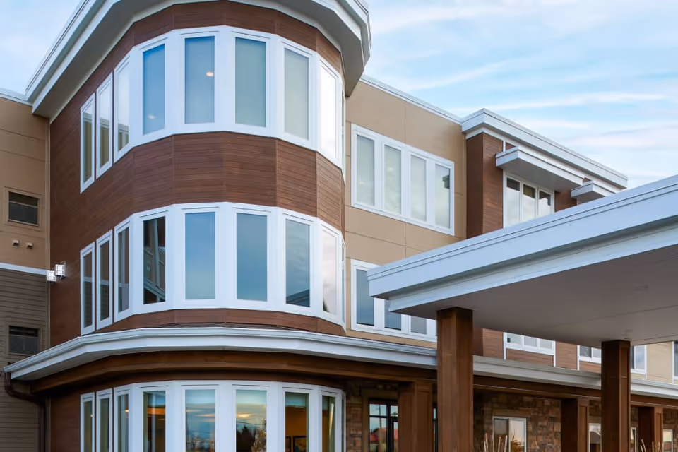 Exterior view of a modern multi-story building with large windows and a covered entrance, featuring brown and beige siding and white trim under a partly cloudy sky.
