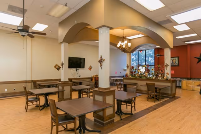 A spacious dining area with several wooden tables and chairs arranged neatly. The room features a light wood floor, beige walls with wooden trim, and a ceiling with recessed lighting and ceiling fans. There is a decorative archway with a chandelier hanging in the center, and a large window letting in natural light. A flat-screen TV is mounted on one wall, and decorative items are placed on the walls and a counter beneath the archway.