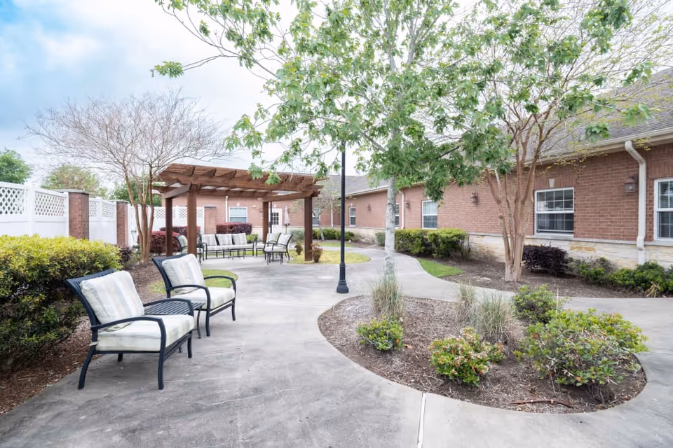 Outdoor courtyard with a paved walkway, cushioned chairs, a wooden pergola, trees and landscaping beside a brick building.