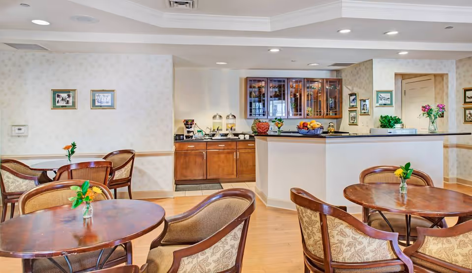 A cozy dining area with round wooden tables and cushioned chairs. Each table has a small vase with a flower. In the background, there is a kitchen counter with cabinets above and below, featuring a coffee station and fruit basket. The walls are decorated with framed pictures and light wallpaper.