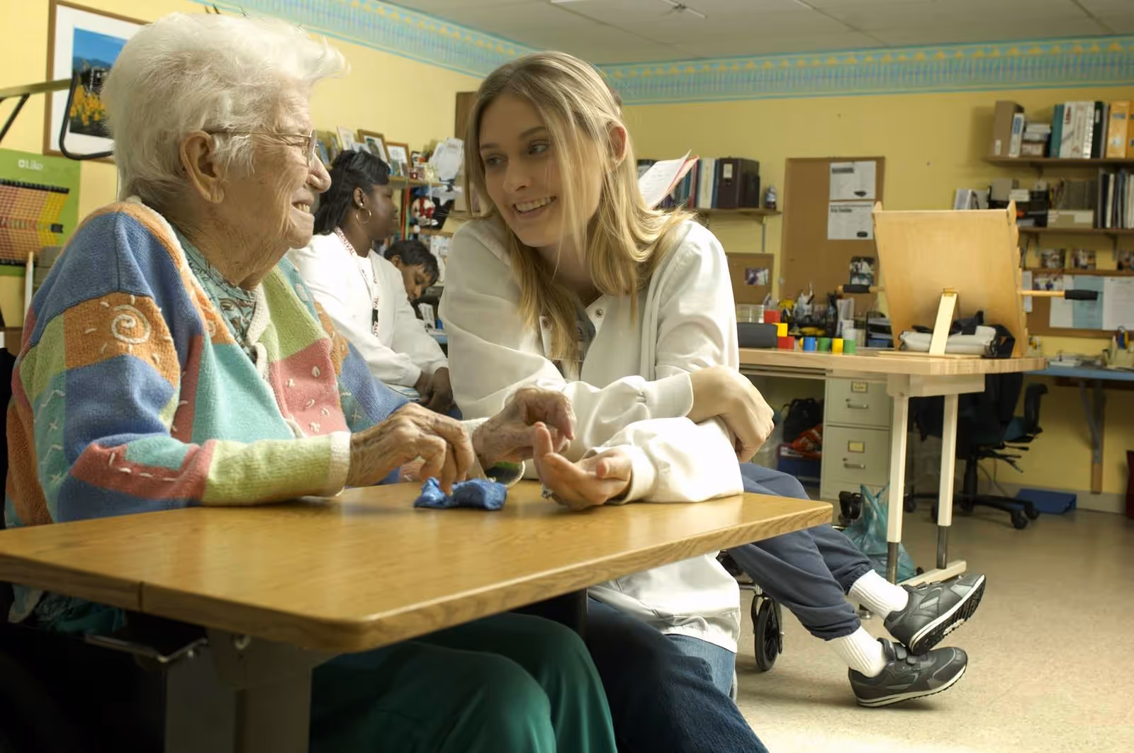 An elderly woman and a young woman sitting at a table in a room that appears to be a recreational or activity area. The elderly woman is wearing a colorful sweater and glasses, and the young woman is smiling and engaging with her. In the background, there are other people and shelves with various items, suggesting a communal or care facility setting.