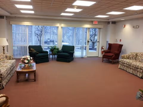 Spacious senior-living lounge with sofas, armchairs, a coffee table, and large glass doors/windows to the outside.