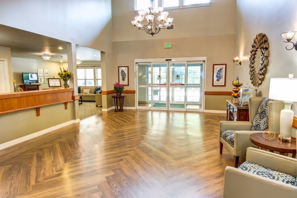 Bright and spacious assisted living facility lobby with wooden herringbone floor, a chandelier hanging from the high ceiling, two patterned armchairs with a small round table and lamp, a decorative mirror on the wall, framed artwork, and glass double doors leading outside.